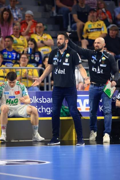 Bennet Wiegert, Cheftrainer des SC Magdeburg, gestikuliert leidenschaftlich an der Seitenlinie während des DAIKIN HBL-Spiels in der SAP Arena. Neben ihm steht ein Betreuer mit Taktikboard, während ein Spieler im weißen SCM-Trikot erschöpft auf der Bank sitzt. SC Magdeburg Trainer Bennet Wiegert gibt Anweisungen an der Seitenlinie in der SAP Arena
