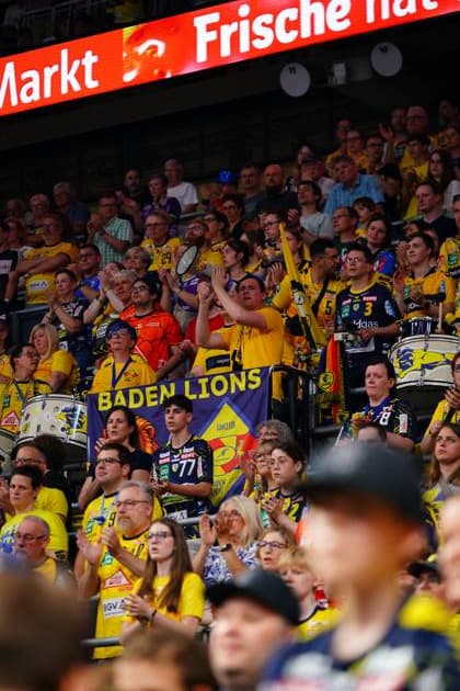 Die Fankurve der Rhein Neckar Löwen ist dicht gefüllt mit Fans in gelben Trikots, die das Spiel verfolgen. Ein lila Banner des Fanclubs Baden Lions ist gut sichtbar im Block, dazu sind Trommeln der Fangruppe zu sehen. Rhein Neckar Löwen Fans mit Baden Lions Banner in der SAP Arena