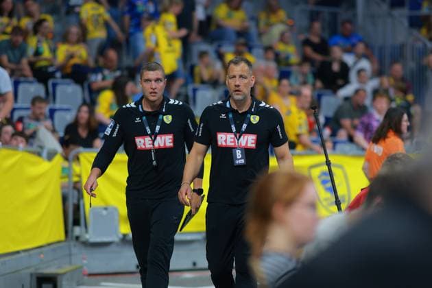 Trainer Maik Machulla und Co-Trainer Michel Abt der Rhein Neckar Löwen laufen in schwarzen Teamoutfits mit REWE-Sponsorenlogo an der Seitenlinie der SAP Arena. Im Hintergrund ist die gelb gekleidete Heimfankurve zu sehen. Maik Machulla und Michel Abt (Rhein Neckar Löwen) an der Seitenlinie