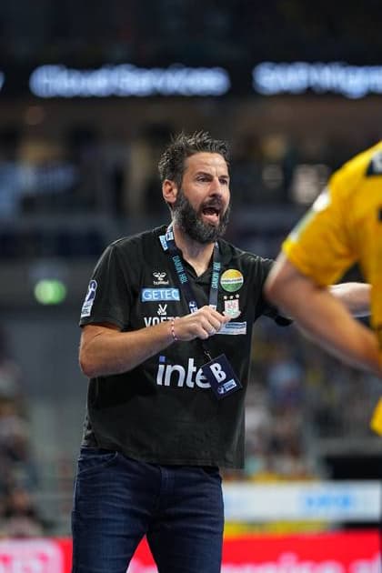 SC Magdeburg Trainer Bennet Wiegert steht mit geöffnetem Mund und gestikulierend an der Seitenlinie in der SAP Arena. Er trägt ein dunkles Trainershirt mit SC Magdeburg-Emblem und einem HBL-Akkreditierungsausweis. Bennet Wiegert (Trainer; SC Magdeburg) gibt Anweisungen an der Seitenlinie