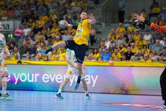 Sebastian Heymann ( 13; Rhein Neckar Löwen) springt in der SAP Arena hoch und zieht zum Wurf ab, während ein SC Magdeburg Torwart im orangefarbenen Trikot den Arm ausstreckt. Im Hintergrund sind SC Magdeburg Spieler in weißen Trikots sowie das vollbesetzte Publikum in der Löwen-Fankurve zu sehen. Sebastian Heymann ( 13; Rhein Neckar Löwen) beim Sprungwurf