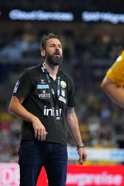 SC-Magdeburg-Trainer Bennet Wiegert steht an der Seitenlinie in der SAP Arena und blickt konzentriert nach oben. Er trägt ein dunkles Trainershirt mit den Logos des SC Magdeburg und der DAIKIN HBL sowie einen Akkreditierungsausweis. Bennet Wiegert (Trainer; SC Magdeburg) verfolgt das Spielgeschehen