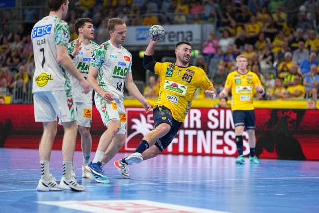 Jannik Kohlbacher ( 80; Rhein Neckar Löwen) springt ab und zieht in gelber Trikotfarbe einen Wurf auf das Tor, während zwei SC Magdeburg-Spieler – darunter Felix Claar ( 7) und Omar Ingi Magnusson ( 14) in weißen Trikots – ihn zu blocken versuchen. Die Szene findet in der vollbesetzten SAP Arena im Rahmen der DAIKIN HBL statt. Jannik Kohlbacher ( 80; Rhein Neckar Löwen) beim Wurfversuch in der SAP Arena
