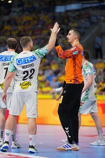 Torwart Matej Mandi? ( 12; SC Magdeburg) im orangenen Torwarttrikot klatscht mit Lukas Mertens ( 22; SC Magdeburg) ab. Im Hintergrund sind weitere Magdeburger Feldspieler sowie die gelb gekleidete Zuschauertribüne der SAP Arena zu sehen. Matej Mandi? ( 12; SC Magdeburg) und Lukas Mertens ( 22) beim High-Five