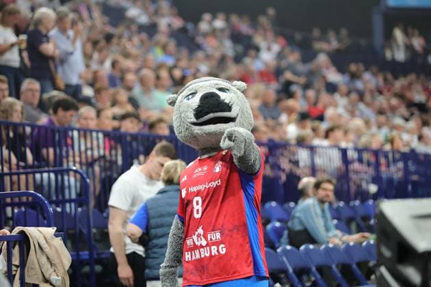 Das Maskottchen des HSV Hamburg in rotem Trikot mit der Nummer 8 präsentiert sich in der Barclays Arena vor vollbesetzten Rängen. Im Hintergrund sind Zuschauer und die Hallenausstattung des Bundesliga-Handballspiels zu sehen. HSV Hamburg Maskottchen bei Barclays Arena Handball Event