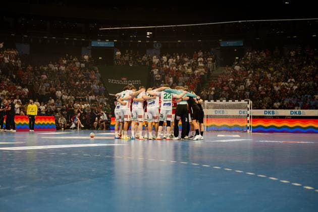 Die Mannschaft von Frisch Auf Göppingen bildet einen geschlossenen Kreis auf dem Spielfeld der Barclays Arena, bevor das Bundesliga-Spiel gegen den HSV Hamburg beginnt. Im Hintergrund füllen Zuschauer die Arena, während Regenbogenflaggen an den Banden sichtbar sind. Frisch Auf Göppingen Team-Huddle vor der Barclays Arena Kulisse Copyright: xPhilippHeistx
