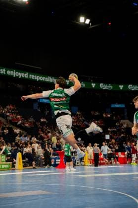 Ein Handballer in grünem Trikot mit Sponsors führt einen Sprungwurf durch und ist dabei vollständig vom Boden abgehoben. Das Spielfeld der Barclays Arena mit Zuschauern im Hintergrund ist gut erkennbar. Spieler in grünem Trikot beim Sprungwurf in der Barclays Arena Copyright: xPhilippHeistx