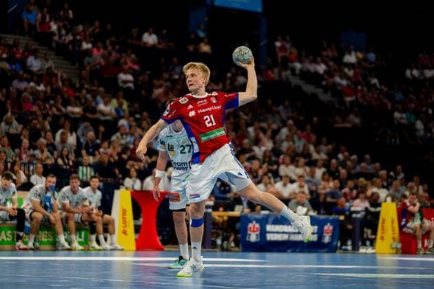 Frederik Bo Andersen trägt das rote Trikot des HSV Hamburg und führt einen Sprungwurf durch, während er den Ball über Kopfhöhe hält. Im Hintergrund sind Spieler von Frisch Auf Göppingen in weißen Trikots und die Zuschauer der Barclays Arena zu sehen. Handball Verein Hamburg vs. Frisch Auf Göppingen Copyright: xPhilippHeistx