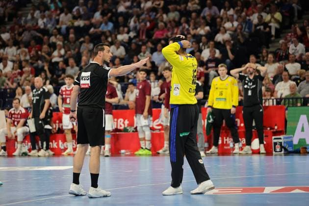 Ein Schiedsrichter in schwarzer Uniform gestikuliert während einer Spielpause. Im Hintergrund stehen Spieler beider Teams auf dem Spielfeld, während Zuschauer in der vollbesetzten Barclays Arena verfolgen. Schiedsrichter bei Spielunterbrechung in der Barclays Arena