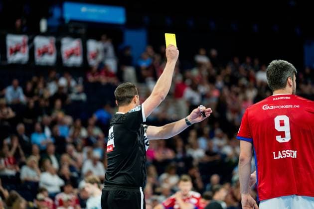 Ein Schiedsrichter hält die gelbe Karte hoch während eines Bundesliga-Spiels zwischen HSV Hamburg und Frisch Auf Göppingen. Im Hintergrund steht Jacob Lassen ( 9; HSV Hamburg) in seinem roten Trikot an der Seitenlinie. Handball Verein Hamburg vs. Frisch Auf Göppingen Copyright: xPhilippHeistx