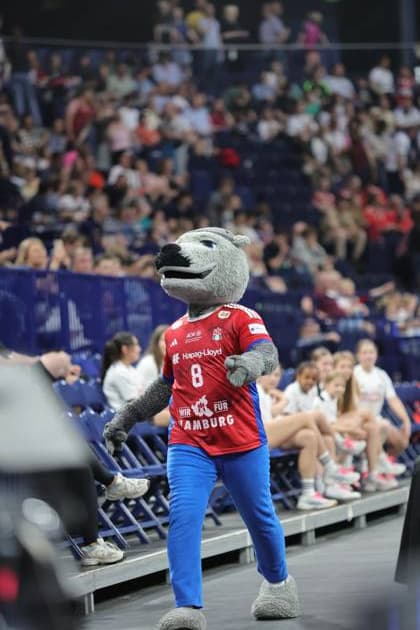 Das HSV-Maskottchen in rotem und blauem Kostüm winkt den Zuschauern in der Barclays Arena zu. Im Hintergrund sind Spieler in blauen HSV-Trikots und Fans in den Rängen zu sehen. HSV Hamburg Maskottchen bei Spielerbetreuung *** The HSV mascot in red and blue costume waves to the spectators in the Barclays Arena In the background you can see players in blue HSV jerseys and fans in the stands HSV Hamburg mascot looking after players Copyright: xspeedshotx