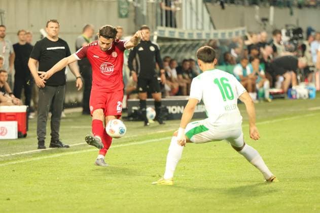 José Enrique Ríos Alonso ( 23; Rot-Weiss Essen) zieht am linken Flügel ab, während Aaron Keller ( 16; SpVgg Greuther Fürth) den Schuss zu blocken versucht. Die Szene findet nahe der Seitenlinie statt, im Hintergrund ist die Trainerbank zu sehen. José Enrique Ríos Alonso ( 23; Rot-Weiss Essen) schießt gegen Aaron Keller ( 16; SpVgg Greuther Fürth)