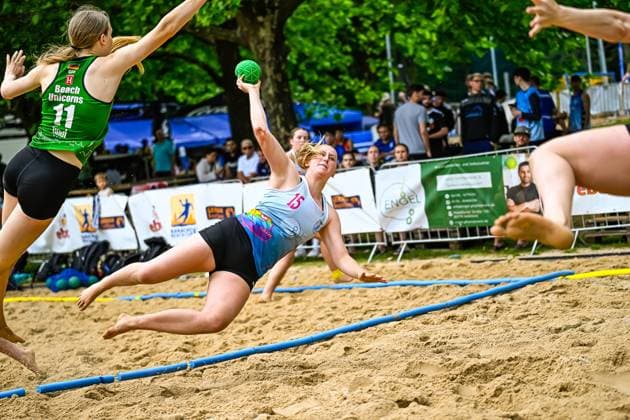 Eine Spielerin ( 15) wirft sich im Flug über den Sand und versucht einen Torwurf, während eine Gegenspielerin der Beach Unicorns ( 11) die Abwehr versucht. Im Hintergrund verfolgen zahlreiche Zuschauer das Spiel. Spektakulärer Wurfversuch ( 15) im Beach-Handball-Spiel gegen Beach Unicorns