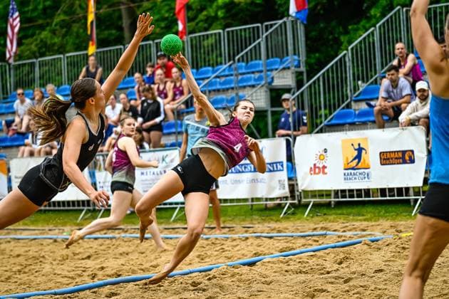 Eine Spielerin im pinken Trikot ( 4) wirft beim German BeachOpen Karacho Beach Cup aus extremer Schräglage auf das Tor, während zwei Gegenspielerinnen in schwarzen Trikots den Abschluss zu blocken versuchen. Im Hintergrund verfolgen Zuschauer das Spiel auf der Tribüne. Spielerin ( 4) beim Wurfversuch beim Karacho Beach Cup