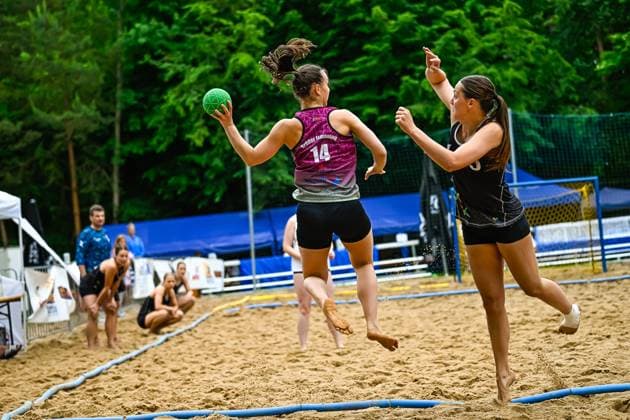 Eine Spielerin von Brüder Ismaning ( 14) springt ab und holt zum Wurf aus, während eine Gegenspielerin im schwarzen Trikot versucht, den Wurf zu blocken. Das Spiel findet auf einem Sandplatz im Freien statt. Spielerin ( 14; Brüder Ismaning) beim Wurfversuch im Beach-Handball