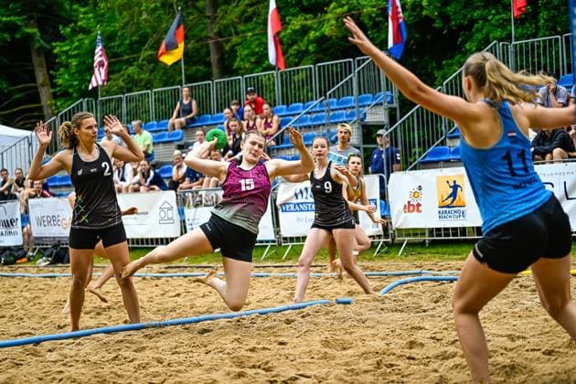 Eine Spielerin mit Trikotnummer 15 springt beim Karacho Beach Cup zum Wurf auf dem Sandfeld. Zwei Spielerinnen mit NED-Aufdruck ( 2 und 9) sowie eine Gegenspielerin ( 11) sind im Bild zu sehen. Spielerin ( 15) beim Wurf beim Karacho Beach Cup
