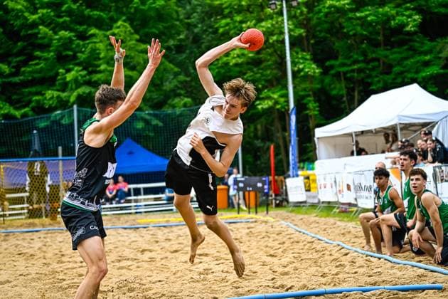 Ein Spieler im weißen Trikot springt hoch und wirft den Ball gegen einen Abwehrspieler im schwarz-grünen Trikot. Im Hintergrund sind weitere Spieler und Zuschauer an der Seitenlinie zu sehen. Wurfaktion beim Beach-Handball: Spieler springt gegen Abwehrblock