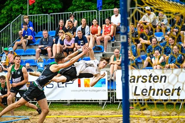 Ein Spieler im weißen Trikot ( 10) wirft sich in einer akrobatischen Flugbewegung zum Tor, während ein Gegenspieler im schwarzen Trikot den Abschluss zu blocken versucht. Die Szene findet auf einem Sandplatz vor gut besetzten Zuschauerrängen statt. Spektakulärer Wurfversuch beim Beach-Handball vor vollem Publikum