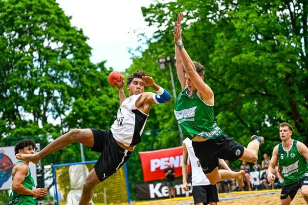Ein Spieler im weißen Trikot springt zum Wurf und wird von einem Gegner in Grün geblockt. Das Spiel findet auf einem Sandplatz im Freien statt, im Hintergrund sind Sponsorenbanner von Kempa und Penny sichtbar. Wurfaktion im Beach-Handball: Spieler im Zweikampf gegen Gegner