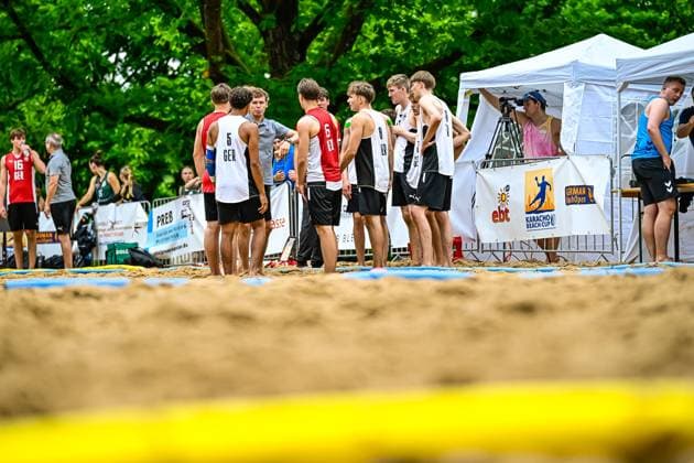 Mehrere Spieler der deutschen Jugend-Nationalmannschaft stehen auf dem Sandfeld beim Karacho Beach Cup zusammen, erkennbar an den Trikots mit GER-Aufdruck und den Nummern 5, 6 und 8. Im Hintergrund sind Werbebanden, ein Kameramann sowie das Turnierzelt zu sehen. Deutschland-Junioren beim Karacho Beach Cup im Teamgespräch