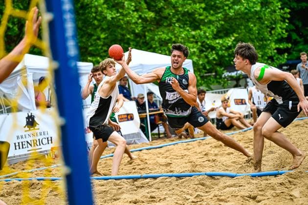 Ein Spieler im grün-schwarzen Trikot mit der Nummer 10 zieht aus dem Rückraum ab und wird dabei von einem Gegenspieler geblockt. Die Szene findet auf einem Sandplatz im Freien statt, im Hintergrund sind Zuschauer und Werbebanden sichtbar. Spieler ( 10) mit Wurfversuch beim Beach-Handball-Turnier