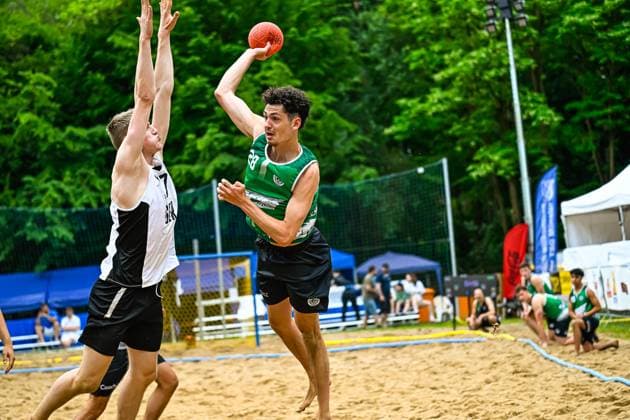 Ein Spieler im grünen Trikot ( 28) springt hoch und wirft den Ball über einen Abwehrspieler im weiß-schwarzen Trikot hinweg. Das Spiel findet auf einem Sandplatz im Freien statt, im Hintergrund sind Zuschauer und Zelte zu sehen. Wurf im Beach-Handball: Grüner Angreifer gegen weiß-schwarze Abwehr