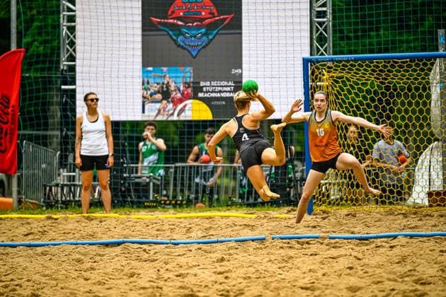 Eine Spielerin mit der Nummer 4 springt in der Luft und wirft den Ball auf das Tor, während Torfrau 16 die Arme ausbreitet und den Wurf abwehren will. Im Hintergrund beobachtet eine Schiedsrichterin die Szene auf dem Sandplatz. Spielerin ( 4) beim Torwurf gegen Torfrau ( 16) im Beachhandball