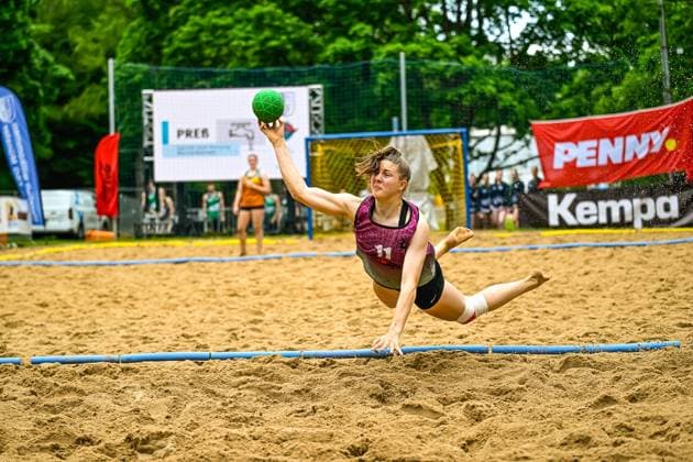 Eine Spielerin mit Trikotnummer 11 wirft im Fallen den Ball auf das Tor und streckt sich dabei horizontal über den Sand. Im Hintergrund sind weitere Spielerinnen sowie Werbebanden von Penny und Kempa zu sehen. Spielerin ( 11) mit Wurfaktion beim Beach-Handball-Turnier