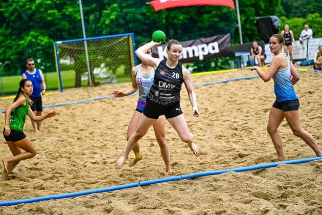 Eine Spielerin mit der Nummer 26 im schwarzen Trikot der DMV Group holt zum Wurf aus und hält den grünen Ball über dem Kopf. Zwei Gegenspielerinnen in hellblauen Trikots versuchen den Wurf zu blocken, während eine weitere Spielerin im grünen Trikot im Hintergrund läuft. Spielerin ( 26; DMV Group) beim Wurfversuch im Beach-Handball