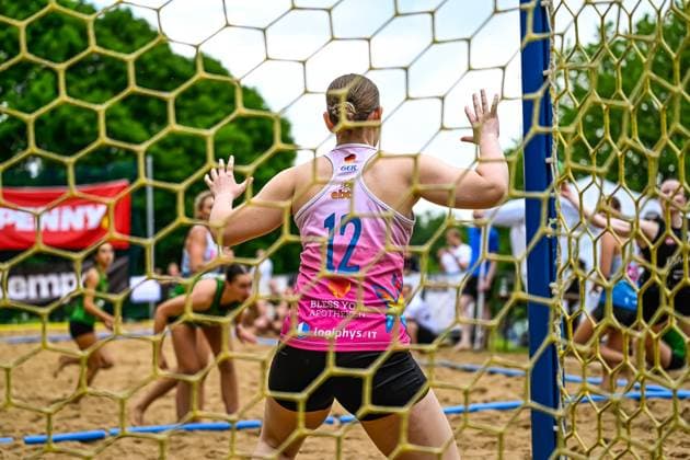 Eine Torfrau im pinken Deutschland-Trikot mit der Nummer 12 steht mit ausgebreiteten Armen im Tor und beobachtet das Spielgeschehen. Das Bild wurde durch das Tornetz aufgenommen, im Hintergrund sind Spielerinnen in grünen Trikots zu sehen. Torfrau 12 (Deutschland) hält das Tor beim Beach-Handball