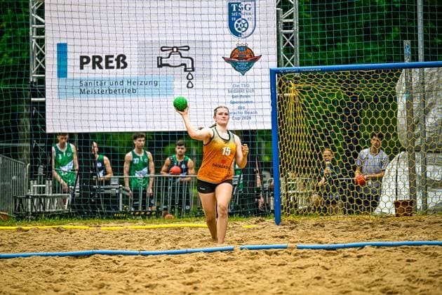 Eine Spielerin mit der Trikotnummer 15 und Aufschrift DATAGROUP holt zum Wurf aus und hält den grünen Handball in der erhobenen Hand. Im Hintergrund sind Spieler in grünen Trikots sowie ein Werbebanner des Aracho Beach Cup der TSG Münster zu sehen. Spielerin ( 15; DATAGROUP) beim Wurf im Beach-Handball