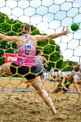 Blick durch das Tornetz auf die deutsche Torfrau ( 12) in pink-weißem Trikot, die mit ausgestrecktem Arm einen grünen Ball abzuwehren versucht. Im Hintergrund sind weitere Spielerinnen im Sand zu sehen. Torfrau 12 (GER) pariert beim Beach-Handball-Turnier