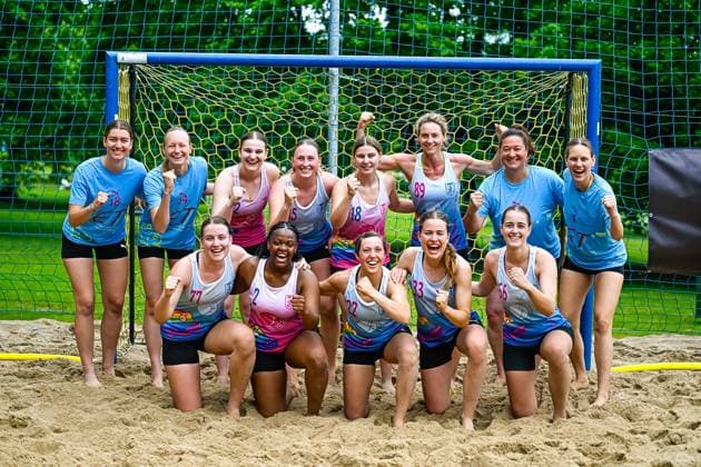 Eine Gruppe von Spielerinnen zweier Teams posiert gemeinsam vor einem blauen Beachhandball-Tor auf Sandboden. Die Spielerinnen zeigen Jubelposen mit geballten Fäusten und lachen in die Kamera. Frauenmannschaft posiert jubelnd vor dem Beachhandball-Tor