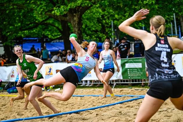 Eine Spielerin mit der Nummer 15 wirft im Sprung auf das Tor, während eine Gegenspielerin der Beach Unicorns ( 4) die Abwehr vorbereitet. Im Hintergrund verfolgt eine Spielerin mit Nummer 12 in grünem Trikot die Szene auf dem Sandplatz. Spielerin ( 15) mit Wurfaktion gegen Beach Unicorns ( 4)