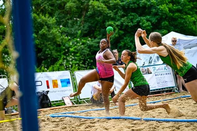 Eine Spielerin im pinken Trikot springt zum Wurf und wird von zwei Gegenspielerinnen in grünen Trikots unter Druck gesetzt. Das Spiel findet auf einem Sandplatz im Freien statt. Wurfaktion beim Beach-Handball-Turnier im Sand