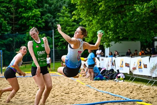 Eine Spielerin im blauen Trikot springt und wirft den Ball in Richtung Tor. Eine Gegenspielerin mit der Nummer 12 im grünen DMV-Group-Trikot beobachtet die Aktion. Wurfaktion beim Beach-Handball-Turnier im Sprung