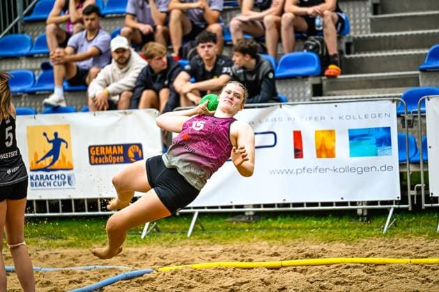 Eine Spielerin im magentafarbenen Trikot wirft sich beim Karacho Beach Cup German Beach Open in einem dynamischen Sprungwurf über den Sandplatz. Im Hintergrund verfolgen Zuschauer auf der Tribüne das Geschehen. Spektakulärer Sprungwurf beim German Beach Open Karacho Beach Cup