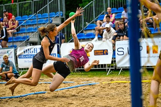 Spielerin 15 im pinken Trikot wirft beim Beachhandball-Turnier German Beach Open in einer spektakulären Flugbewegung auf das Tor, während eine Gegenspielerin im schwarzen Trikot den Block versucht. Der grüne Handball ist im Bild klar zu sehen. Spektakuläre Wurfaktion ( 15) beim German Beach Open