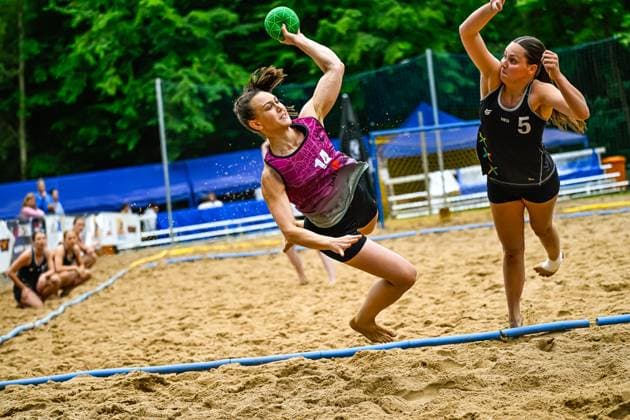 Eine Spielerin mit der Nummer 14 springt dynamisch und wirft den grünen Ball in Richtung Tor, während eine Gegenspielerin der Niederlande ( 5) versucht, den Wurf zu blocken. Das Spiel findet auf einem Sandplatz im Freien statt. Spielerin ( 14) beim Wurfversuch im Beach-Handball-Spiel