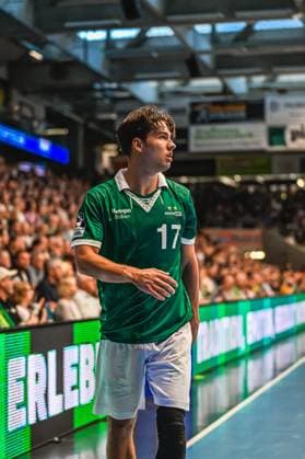 Handball, HBL, Frisch Auf! Göppingen - TVB 1898 Stuttgart #2 Ein Spieler von Frisch Auf! Göppingen mit der Trikotnummer 17 steht an der Seitenlinie und verfolgt konzentriert das Spielgeschehen. Im Hintergrund ist eine gut gefüllte Zuschauertribüne zu sehen. Spieler 17 von Frisch Auf! Göppingen beobachtet das Spielgeschehen