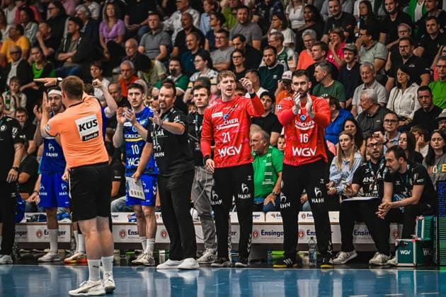 Handball, HBL, Frisch Auf! Göppingen - TVB 1898 Stuttgart #2 Zwei TVB Stuttgart Spieler in roten Trikots ( 21 und 16) gestikulieren emotional an der Seitenlinie neben Betreuern und Trainern. Im Hintergrund ist ein Schiedsrichter im orangenen Trikot sowie ein TVB-Spieler ( 10) in blauem Trikot zu sehen. TVB Stuttgart Spieler ( 21, 16) und Betreuer an der Seitenlinie