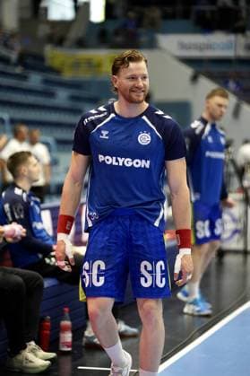 Tom Kiesler ( 30; VfL Gummersbach) läuft lächelnd an der Seitenlinie entlang, mit bandagierten Händen und roten Schweißbändern. Im Hintergrund sind weitere Spieler von VfL Gummersbach sowie die Teambank zu sehen. Tom Kiesler ( 30; VfL Gummersbach) an der Seitenlinie in der SCHWALBE arena