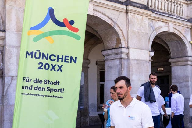 Munich, Bavaria, Germany - June 18, 2026: Poster, banner reading Munich 20XX: For the City and for Sports! next to Sideris Tasiadis (German canoe slalom athlete and Olympian, police officer). press conference, PK, Pressekonferenz held by the top sports federations in front of Nymphenburg Palace to present Munich™s expanded bid concept, as well as the unique features and opportunities the state capital offers as a national candidate for the Summer Olympic Games, Olympische Spiele, Olympia, OS