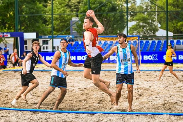 Ein Spieler im roten Trikot springt zum Wurf und hält den Ball über dem Kopf, umgeben von argentinischen Gegenspielern im hellblau-weißen Trikot mit ENARD-Sponsoring. Im Hintergrund ist ein Spieler in schwarzem Trikot zu sehen, der dem deutschen Team zuzuordnen ist. IHF Beach Handball World Championship, WM, Weltmeisterschaft 2026, 23.06.2026, Zagreb, Jarun See *** A player in a red jersey jumps to shoot and holds the ball above his head, surrounded by Argentine opponents in light blue and white jerseys sponsored by ENARD. In the background, a player in a black jersey can be seen; he is part of the German team. IHF Beach Handball World Championship 2026, June 23, 2026, Zagreb, Lake Jarun,detected_source_language Copyright: xspeedshotx xDorianfrickx