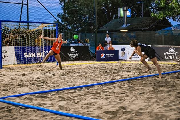 Joelle Arno ( 2; Deutsche Frauen Beachhandball Nationalmannschaft) streckt sich im orangefarbenen Torwart-Trikot weit aus, um einen anfliegenden Ball zu parieren. Eine Feldspielerin in schwarzem Trikot läuft auf das Tor zu, der Ball ist deutlich im Bild sichtbar. IHF Beach Handball World Championship, WM, Weltmeisterschaft 2026, 23.06.2026, Zagreb, Jarun See *** Joelle Arno 2 Copyright: xspeedshotx xDorianfrickx