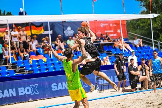 Ein Spieler der deutschen Beachhandball-Nationalmannschaft springt hoch und holt zum Wurf aus, während ein brasilianischer Gegenspieler ( 14) mit erhobenen Armen blockt. Die Szene findet am Jarun See in Zagreb vor einer gut besetzten Tribüne mit deutschen Fahnen statt. IHF Beach Handball World Championship, WM, Weltmeisterschaft 2026, 23.06.2026, Zagreb, Jarun See *** A player on the German national beach handball team jumps high and winds up to shoot, while a Brazilian opponent (No. 14) blocks the shot with his arms raised. The scene takes place at Lake Jarun in Zagreb in front of a well-attended crowd waving German flags. IHF Beach Handball World Championship 2026, June 23, 2026, Zagreb, Lake Jarun,detected_source_language Copyright: xspeedshotx xDorianfrickx