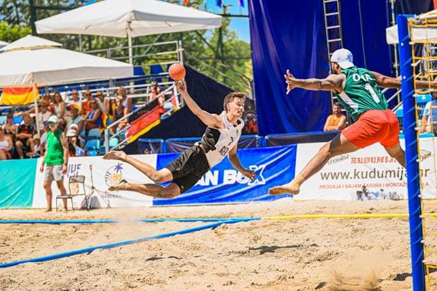 Ein Spieler der deutschen Beachhandball-Nationalmannschaft wirft sich in einer akrobatischen Flugbewegung mit dem Ball in der Hand über den Sand, während ein Gegenspieler mit der Nummer 1 am Tor versucht, den Wurf zu blocken. Die Szene findet am Jarun See in Zagreb vor einer gut besuchten Tribüne statt. IHF Beach Handball World Championship, WM, Weltmeisterschaft 2026, 23.06.2026, Zagreb, Jarun See *** A player on the German national beach handball team leaps across the sand in an acrobatic move, the ball in his hand, while an opposing player wearing number 1 at the goal tries to block the shot. The scene takes place at Lake Jarun in Zagreb in front of a well-attended crowd. IHF Beach Handball World Championship 2026, June 23, 2026, Zagreb, Lake Jarun Copyright: xspeedshotx xDorianfrickx