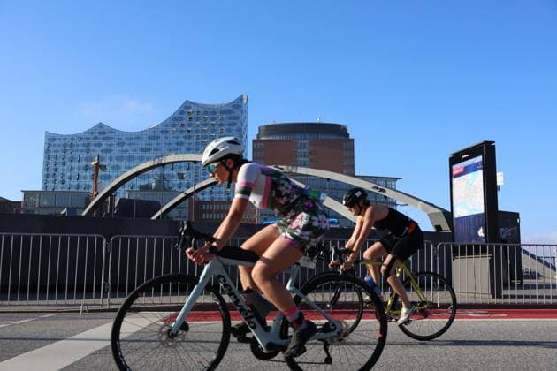 Zwei Athletinnen absolvieren den Radabschnitt des Triathlons am Jungfernstieg in Hamburg, im Hintergrund ist die Elbphilharmonie deutlich zu erkennen. Die vordere Fahrerin trägt ein geblümtes Trikot und einen weißen Helm, die hintere ein schwarzes Outfit mit gelbem Rennrad. Zwei Triathletinnen auf der Radstrecke vor der Elbphilharmonie in Hamburg *** Two athletes are completing the cycling leg of the triathlon on Jungfernstieg in Hamburg, with the Elbphilharmonie clearly visible in the background. The rider in front is wearing a floral jersey and a white helmet; the one behind is wearing a black outfit with a yellow road bike. Two triathletes on the cycling course in front of the Elbphilharmonie in Hamburg Copyright: xspeedshotx xSarahxWolfx