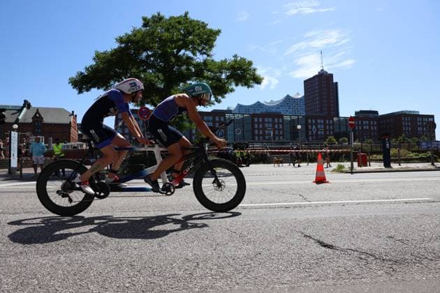 Zwei Athleten auf einem Tandem-Fahrrad während der Radstrecke des Triathlons an der Binnenalster. Die Hamburger Hafenkulisse mit charakteristischen Gebäuden bildet die Kulisse. Tandem-Duo beim Triathlon am Jungfernstieg in Hamburg *** Two athletes on a tandem bike during the cycling leg of the triathlon on the Binnenalster. The Hamburg harbor, with its distinctive buildings, serves as the backdrop for the tandem duo at the triathlon on Jungfernstieg in Hamburg Copyright: xspeedshotx xSarahxWolfx