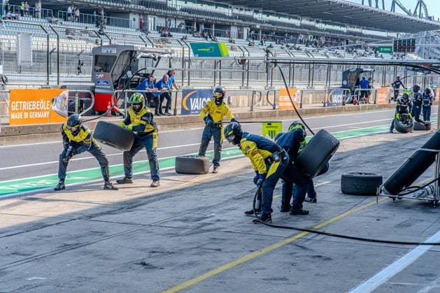 Mehrere Mechaniker in gelb-grauen Overalls üben einen koordinierten Reifenwechsel in der Boxengasse des Nürburgrings. Im Hintergrund sind Werbebanden von VAICO und Getriebeöle sowie ein Ford Mustang GT3-Fahrzeug der HRT Ford Racing zu erkennen. Boxenstopp-Training im Nürburgring Fahrerlager – Mechaniker üben Reifenwechsel *** Several mechanics in yellow-and-gray coveralls are practicing a coordinated tire change in the Nürburgring pit lane. In the background, advertising banners for VAICO and Getriebeöle can be seen, as well as an HRT Ford Racing Ford Mustang GT3. Pit stop training in the Nürburgring paddock – Mechanics practice tire changes Copyright: xMichaelxSchäferx